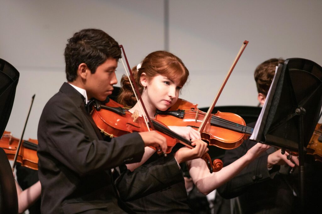 Two violinists playing their instruments and looking at sheet music on a stand, off camera. One violinist appears male with darker skin and black har, wearing a black suite. One violinist appears female with lighter skin and longer red hair, pulled back, wearing a black dress.